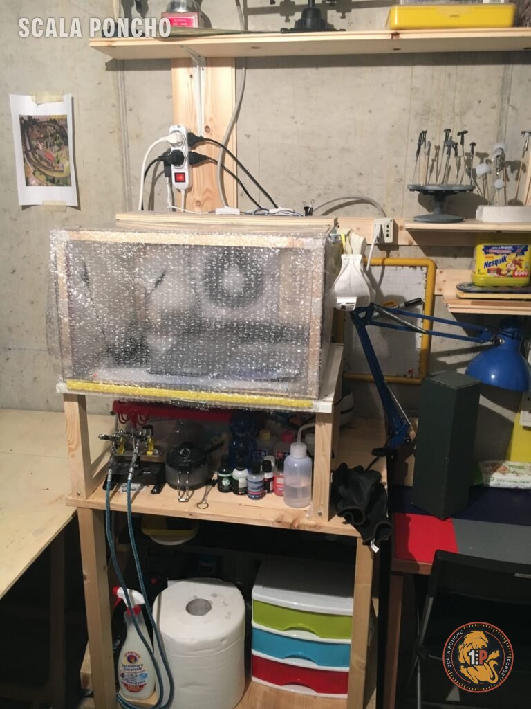 Cabina di verniciatura chiusa con pluriball sopra il banco da lavoro, aerografi e colori Tamiya sul ripiano sotto